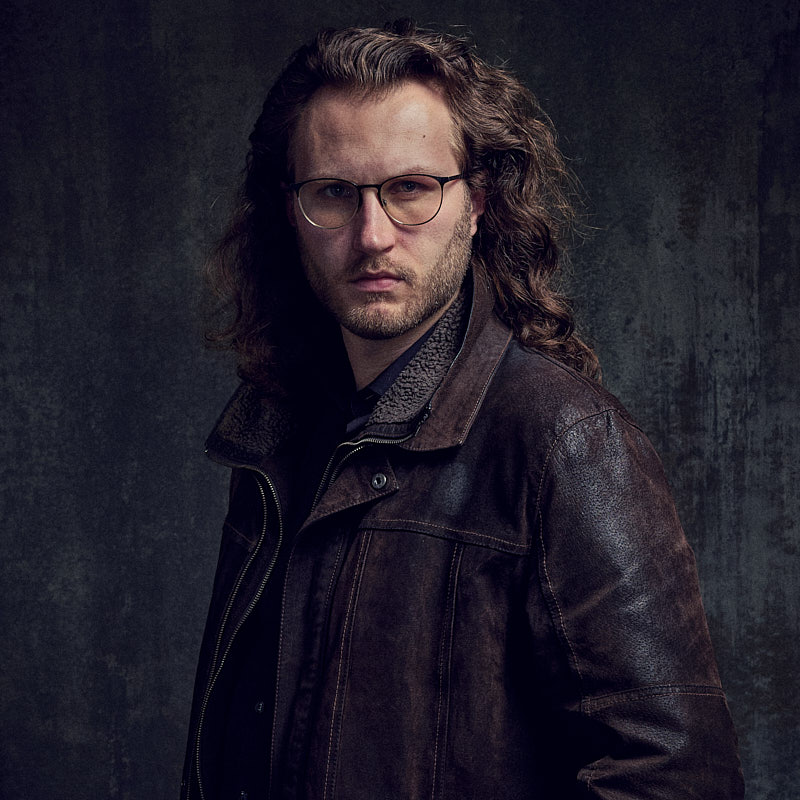 Charakterstarkes Schauspielerportrait eines Mannes mit Brille und brauner Lederjacke vor strukturiertem Hintergrund, erstellt vom Experten in Freising.